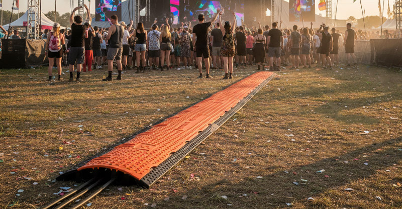 Een kabelbeschermer van Orange Cross 3 ligt op het gras tijdens een festival, voor een publiek dat naar het podium kijkt.