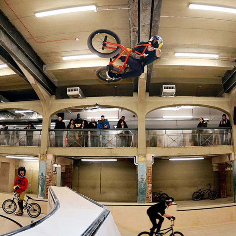 Zdjęcie zrobione w Source BMX Skate Park, przedstawiające osobę w powietrzu na rowerze, obserwowaną przez innych ludzi.
